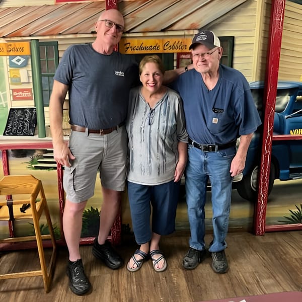 Three smiling people standing together in front of a painted backdrop of a rustic building.
