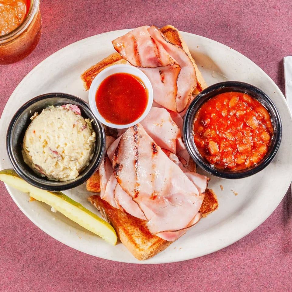 Grilled ham sandwich with potato salad, baked beans, a pickle spear, and BBQ sauce.