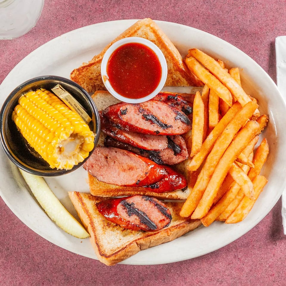 Grilled sausage slices on toast with fries, corn on the cob, a pickle, and BBQ sauce.